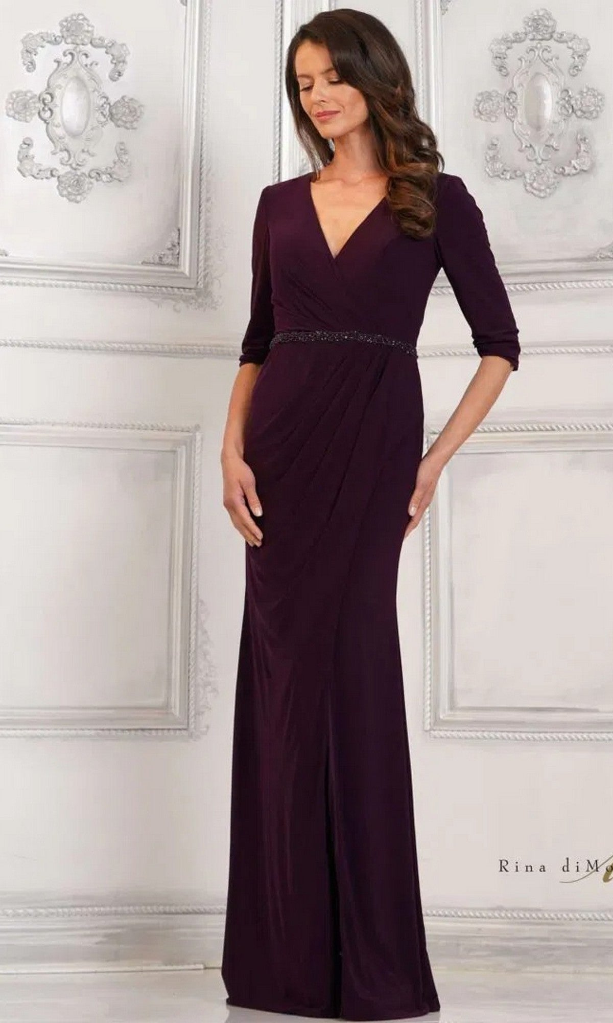 A woman stands against an ornate white wall wearing the Rina Di Montella RD3109 long formal dress in aubergine, featuring a sheath silhouette and three-quarter sleeves.